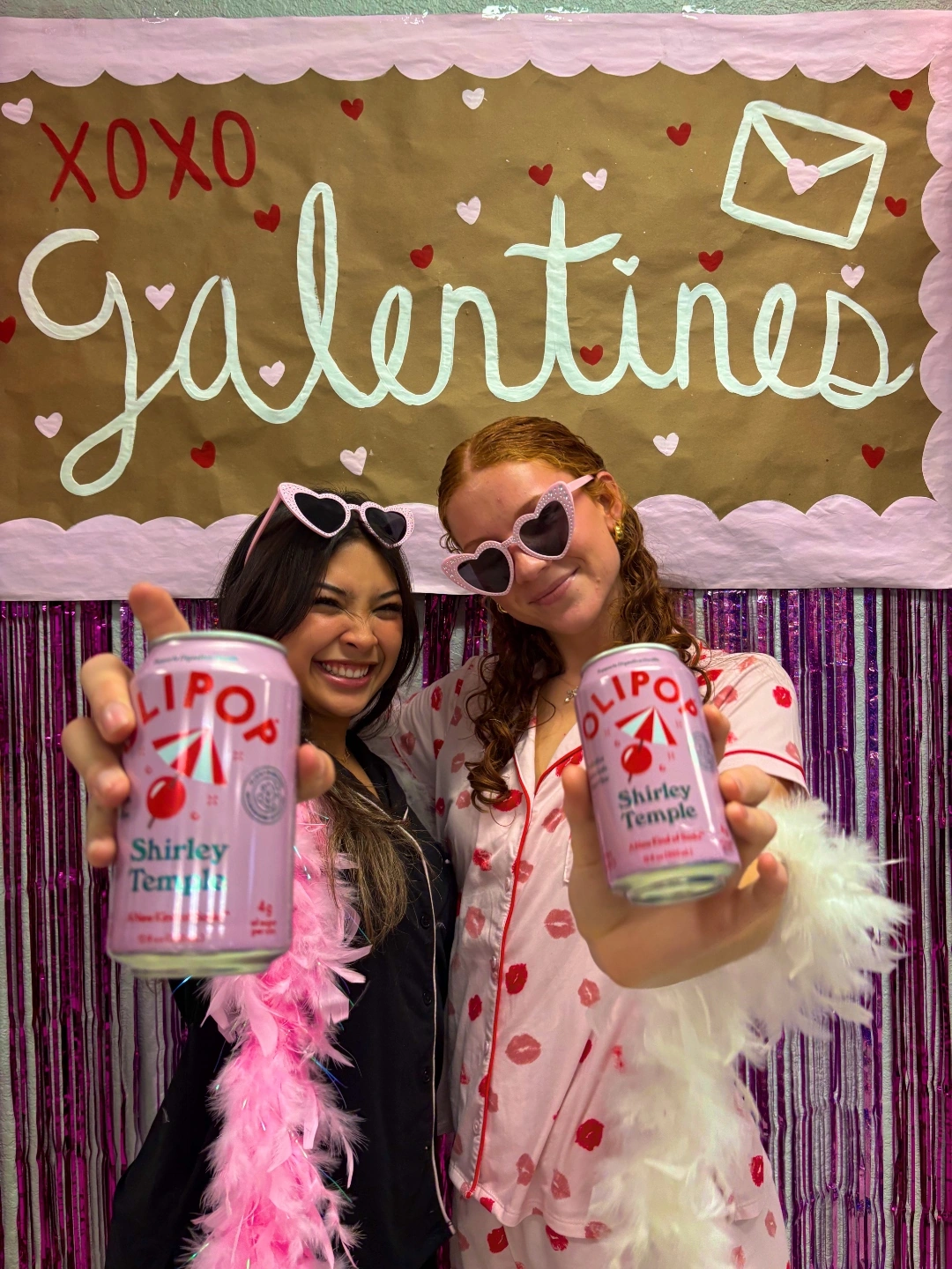 Two college students at a sorority Galentine's event holding OLIPOP Shirley Temple cans in pajamas and feather boas